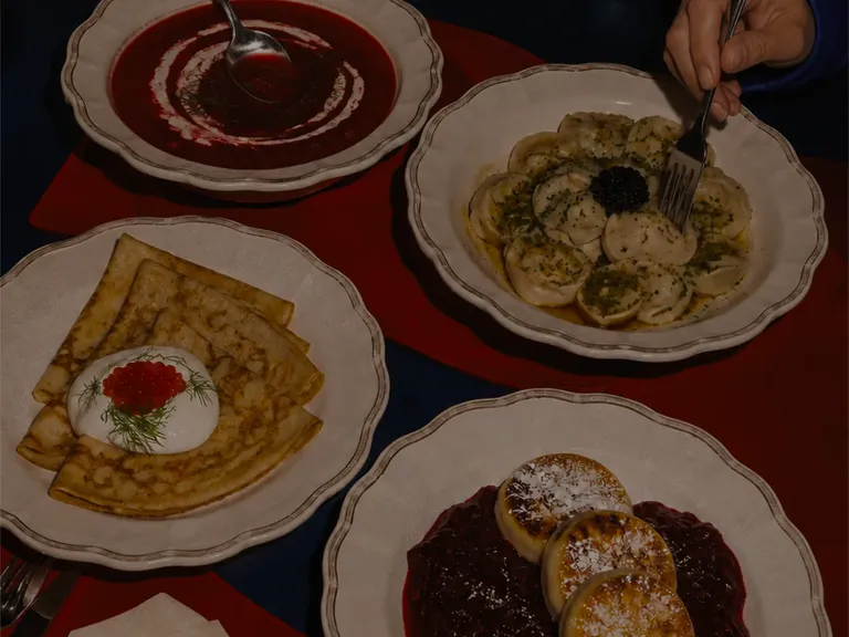 Overhead shot of borscht, pelmeni with caviar, blini with sour cream and roe, and syrniki with berry compote, shared at the table. Client-provided photography.