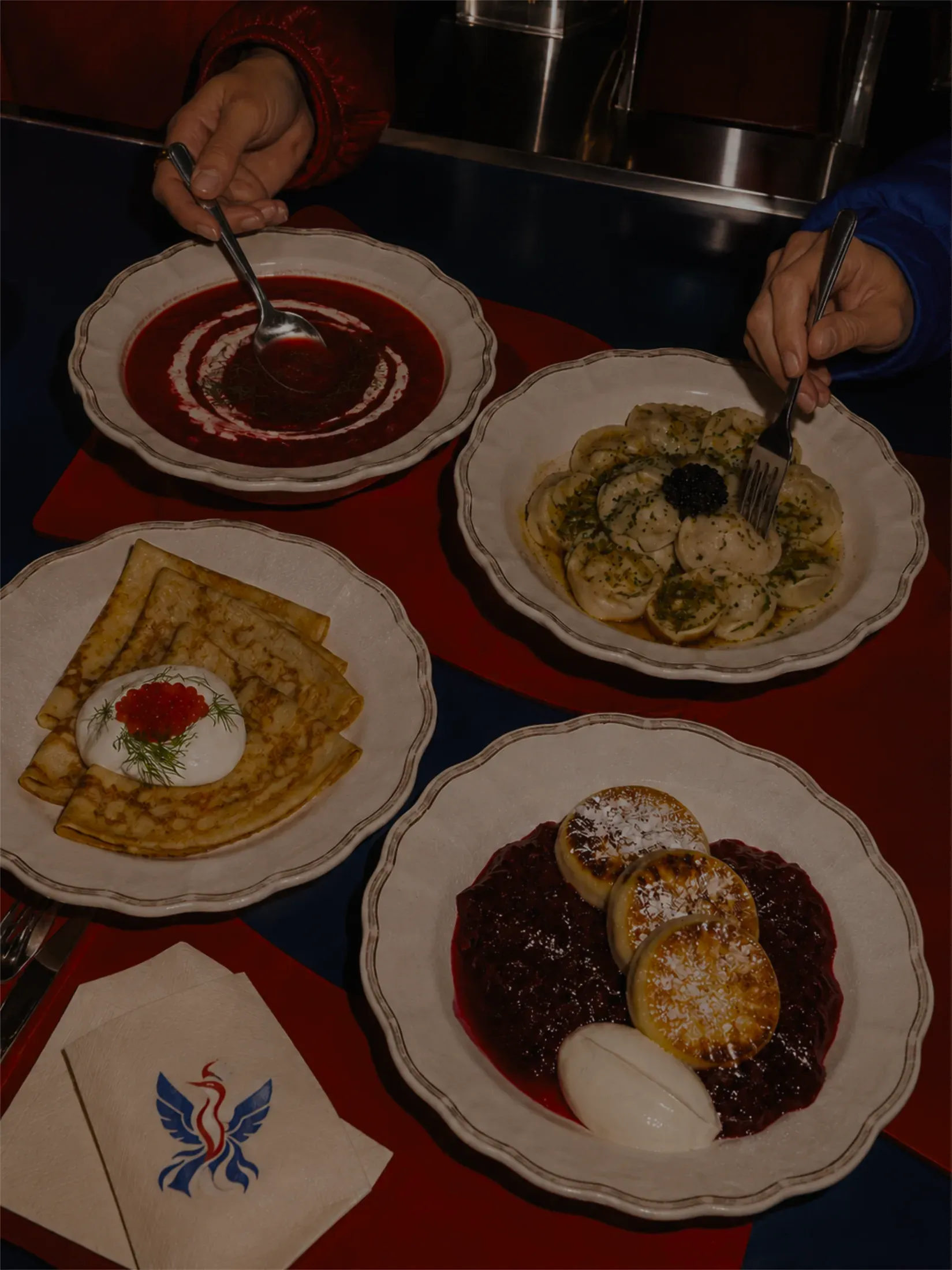 Overhead shot of borscht, pelmeni with caviar, blini with sour cream and roe, and syrniki with berry compote, shared at the table. Client-provided photography.