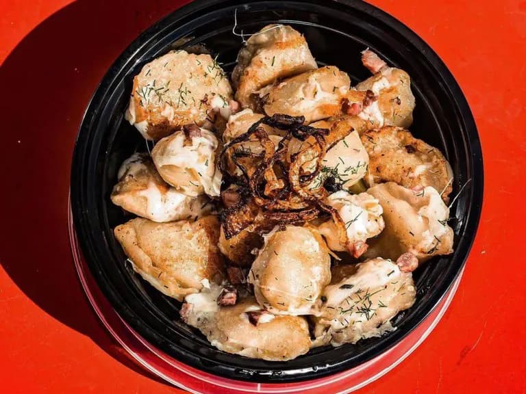 Pierogi in a takeout container with crispy onions, dill and sour cream, on a red table. Client-provided photography.