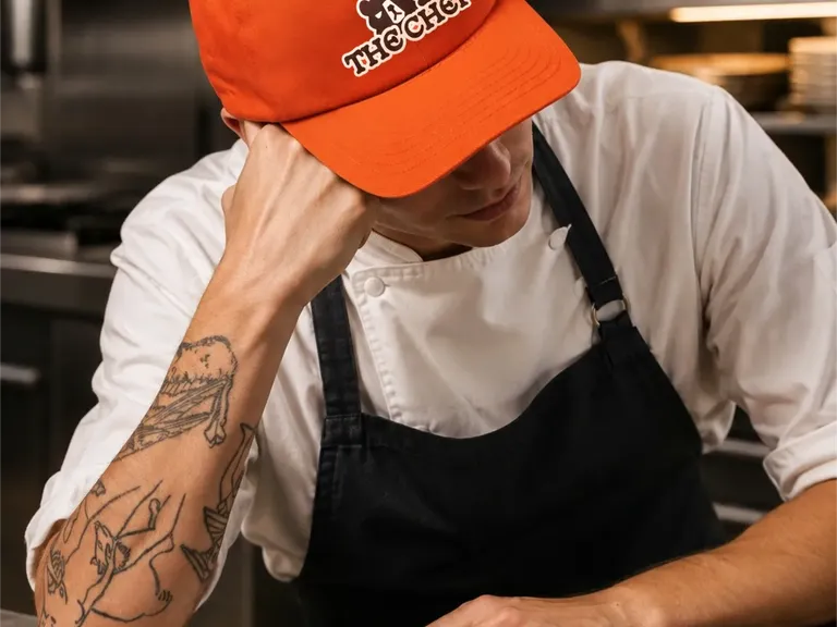 A chef in an orange Borscht Bureau cap embroidered with the bear mark, leaning on a kitchen pass.
