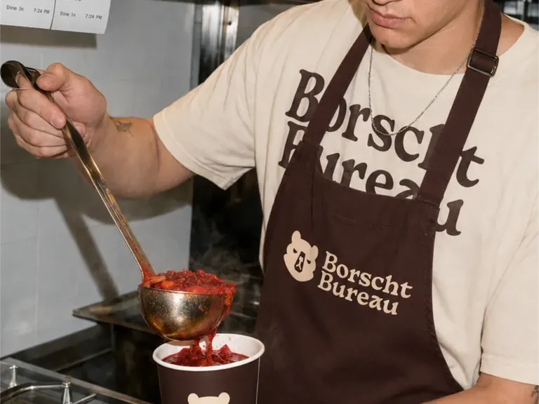 Counter staff in a Borscht Bureau apron and tee ladling borscht into a branded takeaway cup, stacked cups and boxes behind.