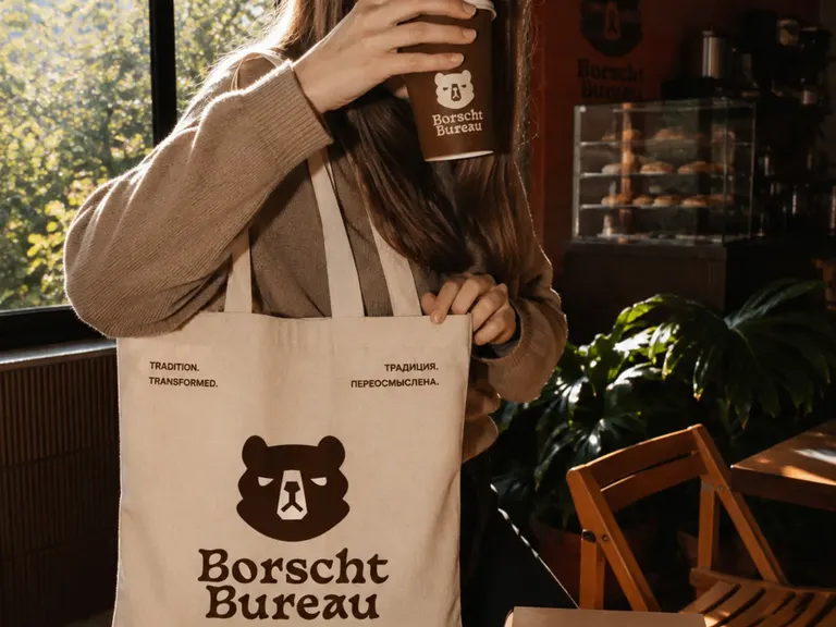 Canvas Borscht Bureau tote bag with the bear mark, held beside a branded coffee cup and pastry box.
