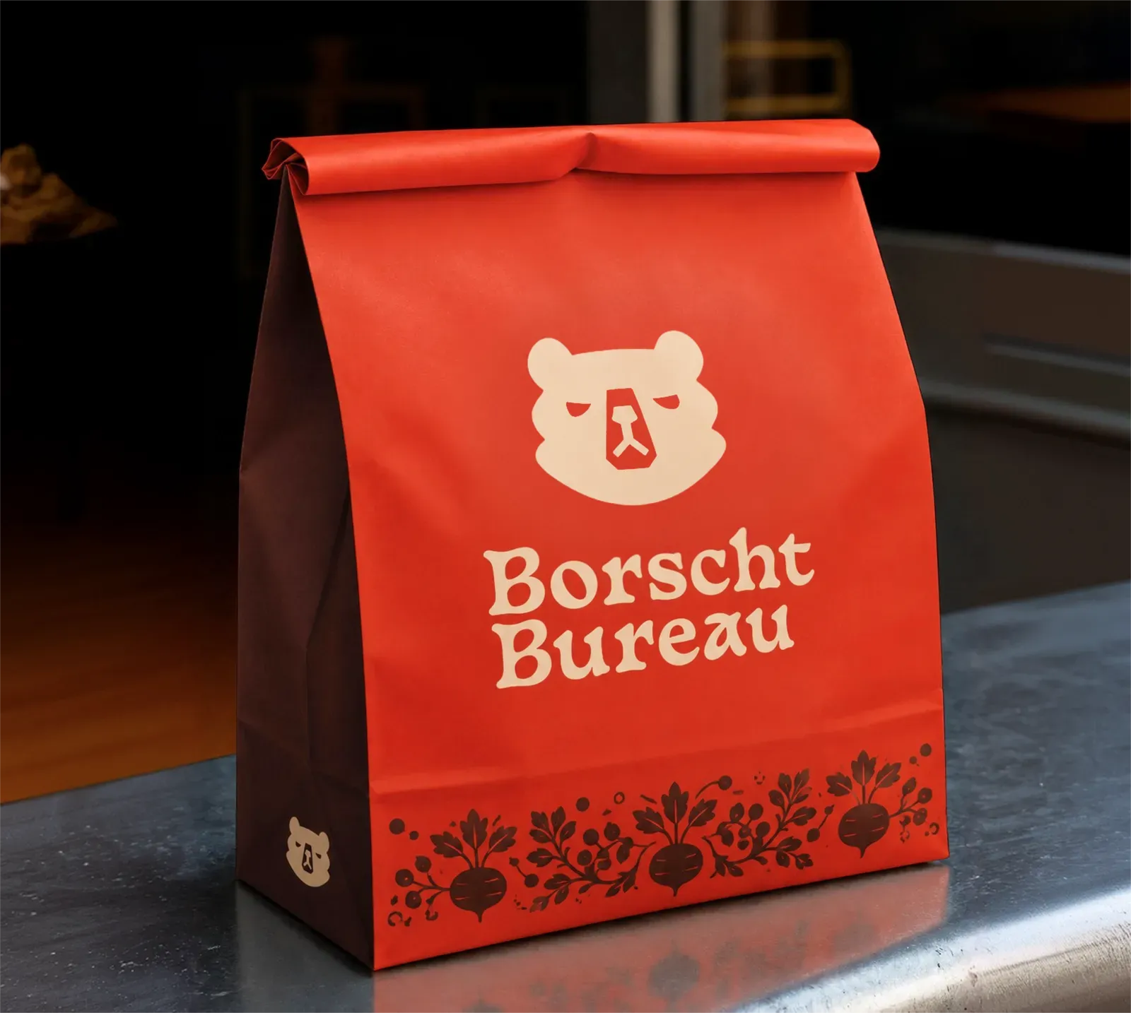 A red Borscht Bureau takeaway bag with the bear mark and the folk beet border printed along the base.